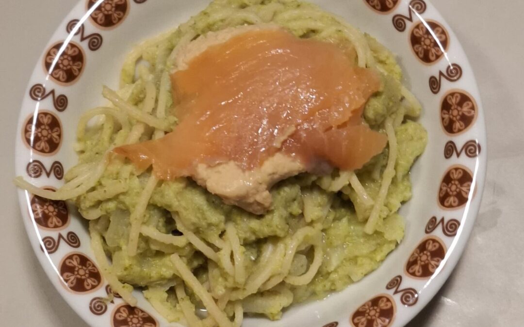 Piatto di spaghetti integrali conditi con crema di broccoli e hummus, guarniti con una fetta di salmone affumicato, serviti in un piatto decorato vintage.